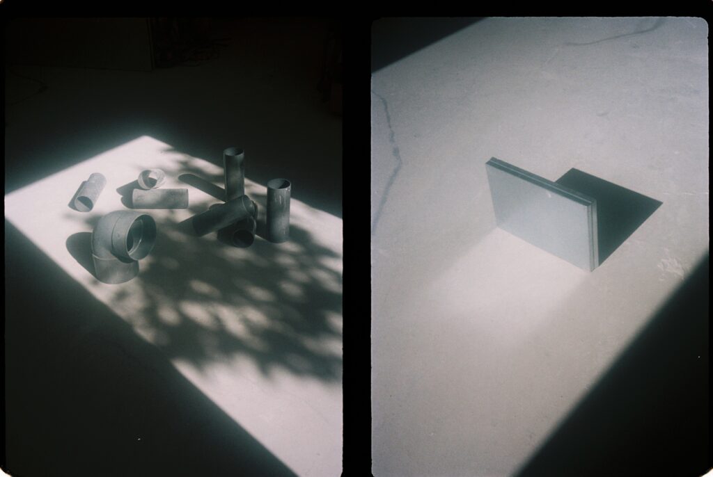 Restes de tub de PVC disposades com un bodegó sota l'ombra d'un arbre, i una peça de fusta recoberta d'inoxidable al mateix lloc
